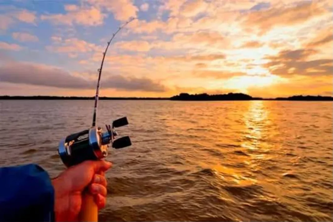 Una mano sostiene un reel de pesca frente a un río al atardecer.